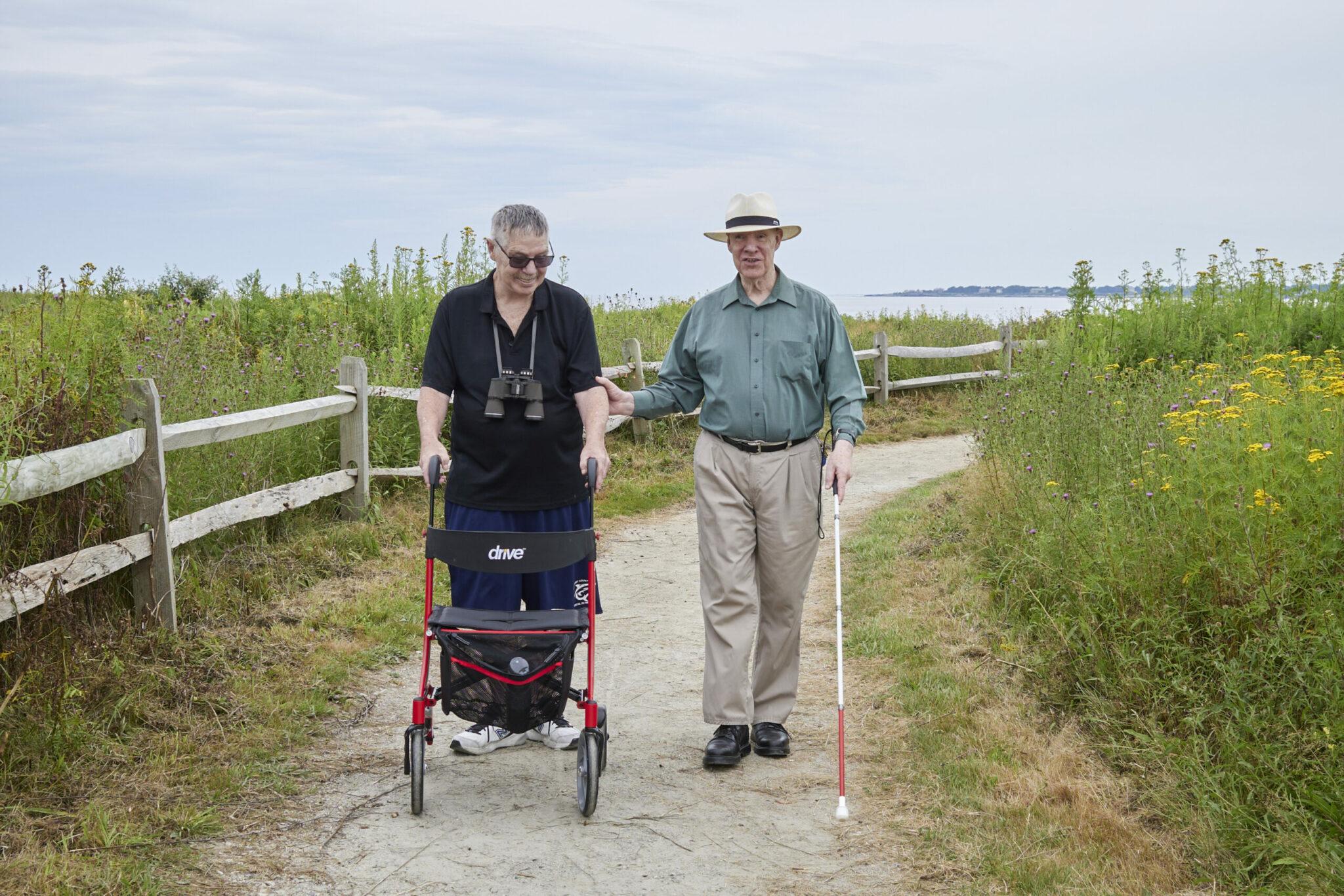 Two older men walking together on a scenic path. One man, wearing a black shirt and binoculars around his neck, uses a red walker, while the other man, dressed in a green shirt and a hat, uses a white cane for assistance. They are surrounded by lush greenery and wildflowers, with a wooden fence lining the path and water visible in the distance. The mood is peaceful and supportive as they enjoy their outdoor walk.