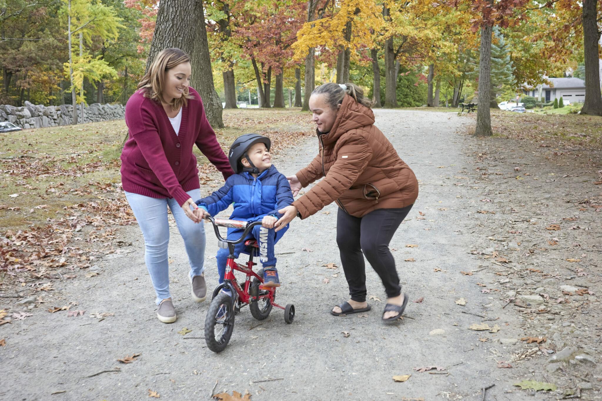 Mother helps son learn to ride bike assisted by smiling staff person