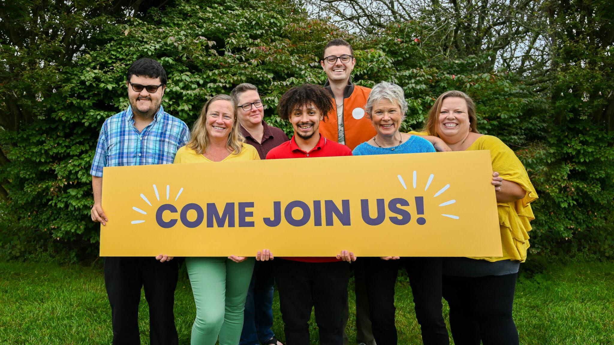 Group of smiling people holding bright yellow sign that says ‘Come Join Us’