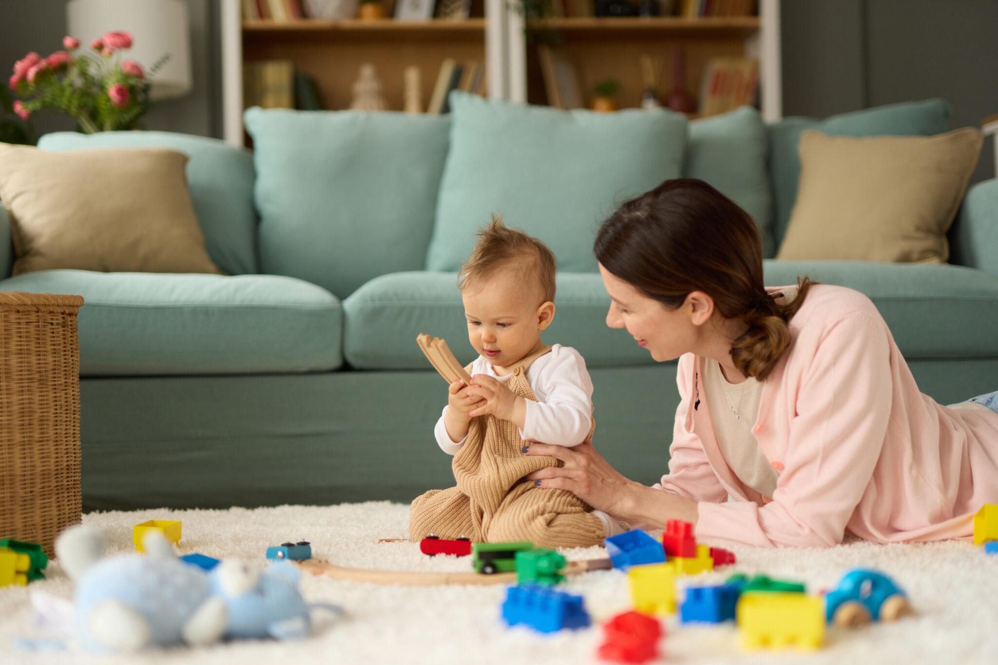 A mother playing with her baby on a soft white carpet surrounded by colorful toys, enjoying quality time in a cozy living room with a light blue couch in the background.