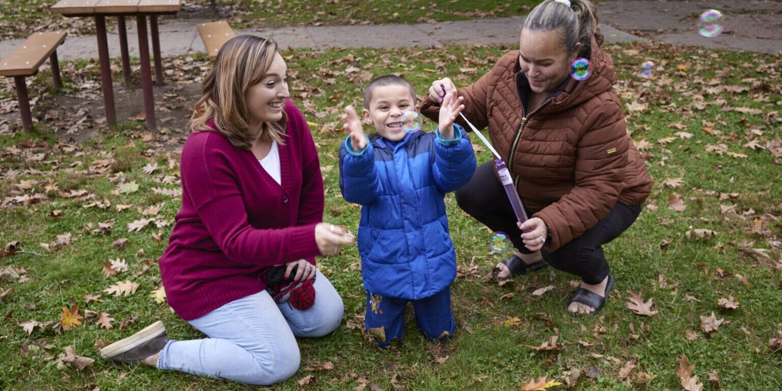 Mother and son in park playing with bubbles accompanied by young woman staff person