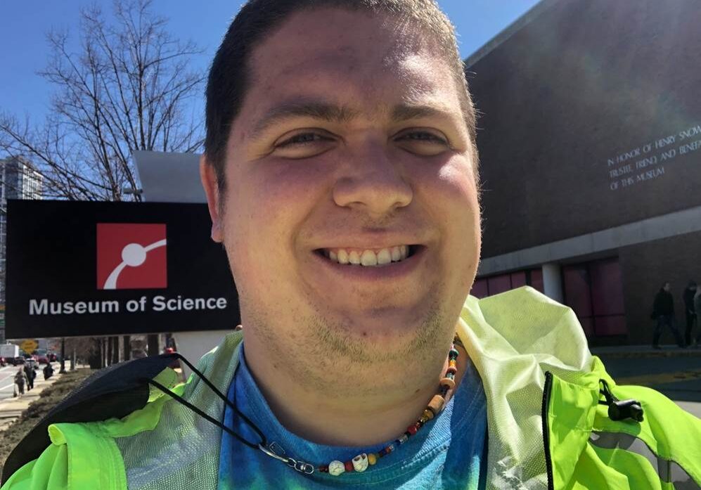 A smiling man takes a selfie in front of the Museum of Science, wearing a bright green jacket and a colorful beaded necklace. The museum’s sign is visible in the background, along with a clear blue sky and bare tree branches, indicating a sunny day.