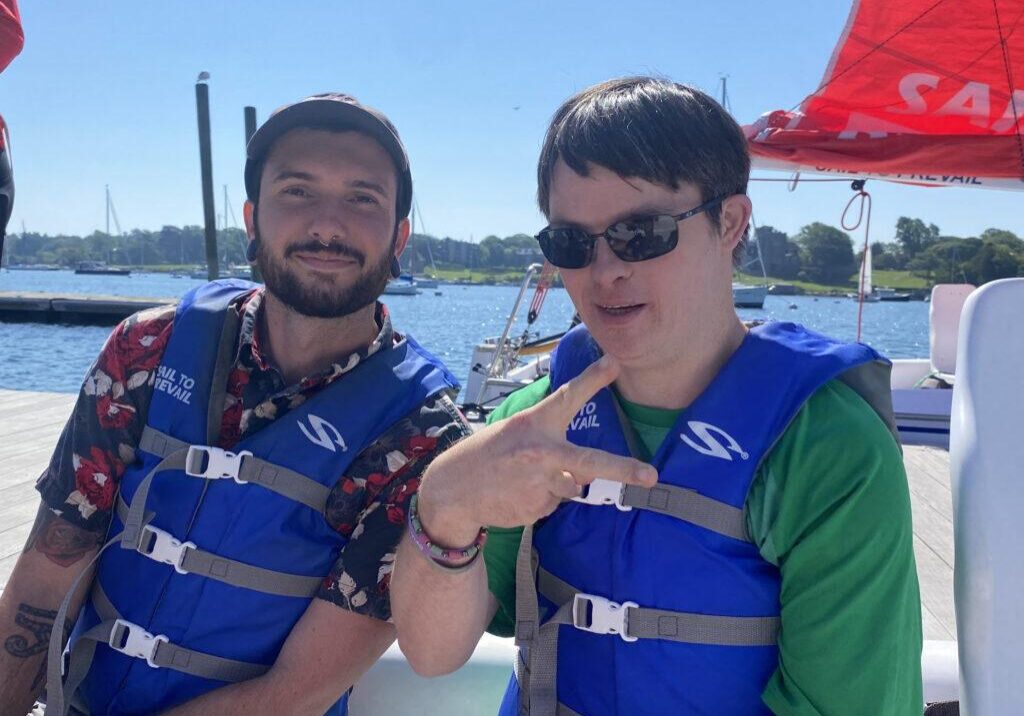 Two men wearing blue life vests are sitting on a dock by the water, ready to sail. The man on the left has a beard and is smiling at the camera, while the man on the right, wearing sunglasses, is making a peace sign with his fingers. In the background, sailboats are visible on a sunny day with clear blue skies.